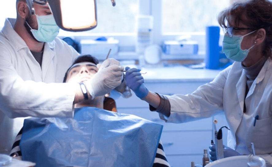 A dentist and his assistant, both wearing masks and gloves, perform an oral surgery procedure on a patient in a dental chair under bright clinical lighting.